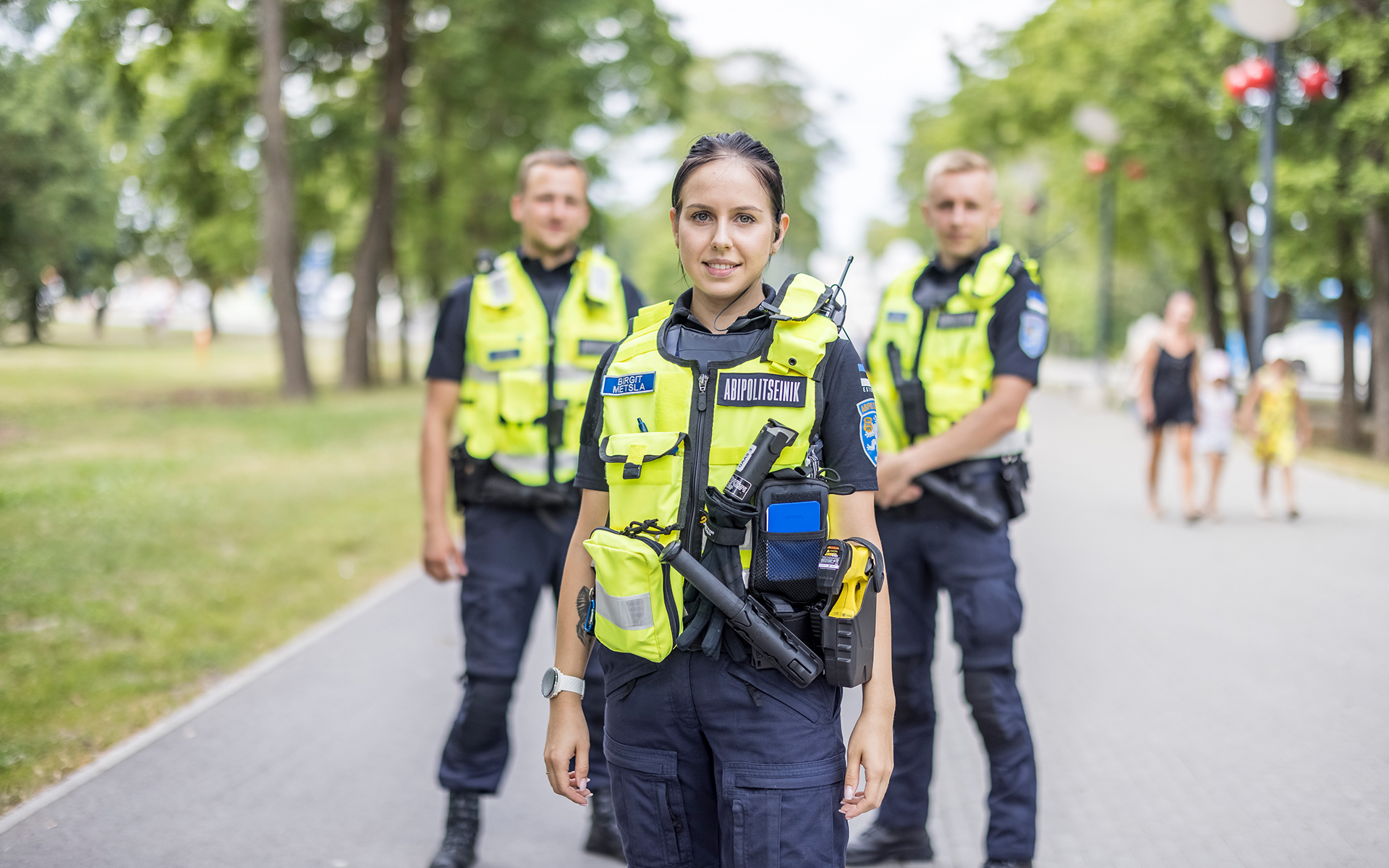 Esiplaanil kollases vestis nais-abipolitseinik, tagaplaanil kaks samuti kollastes vestides meest. Kõik kolm seisavad laial pargiteel, mõlemal pool teed paistavas taamal puud, politseinikest paremal pere lastega. 